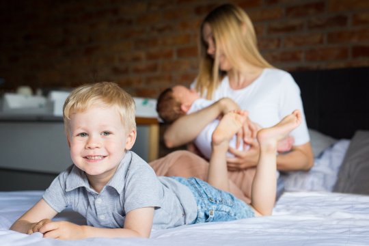 chłopiec podczas sesji zdjęciowej w domu leży na łóżku i uśmiecha się do zdjęcia w tle jest mama z bratem